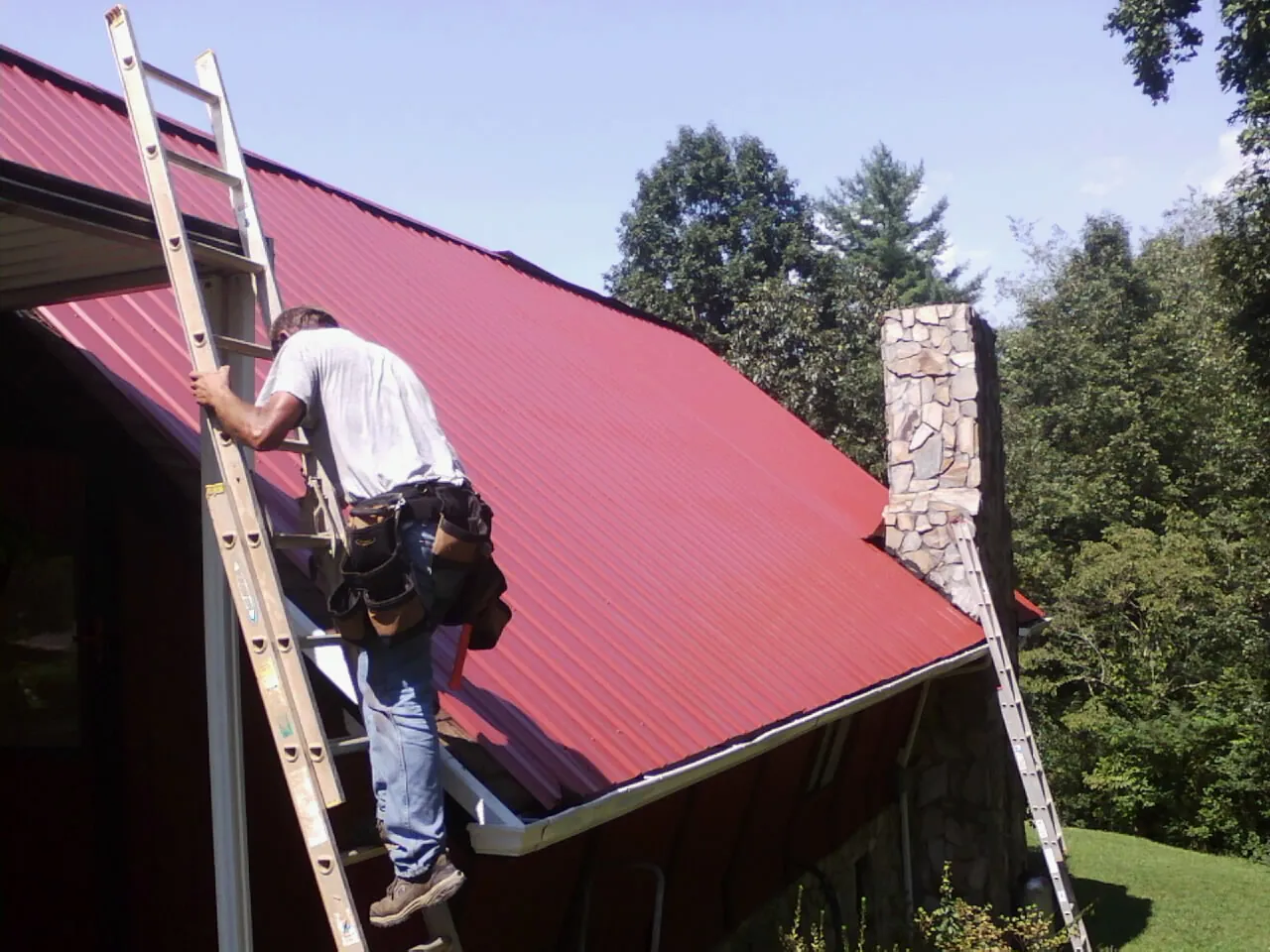 Roofer on a ladder beside a red metal roof and stone chimney — Soffit & Fascia Repair by Roofing Brothers in Millers Falls